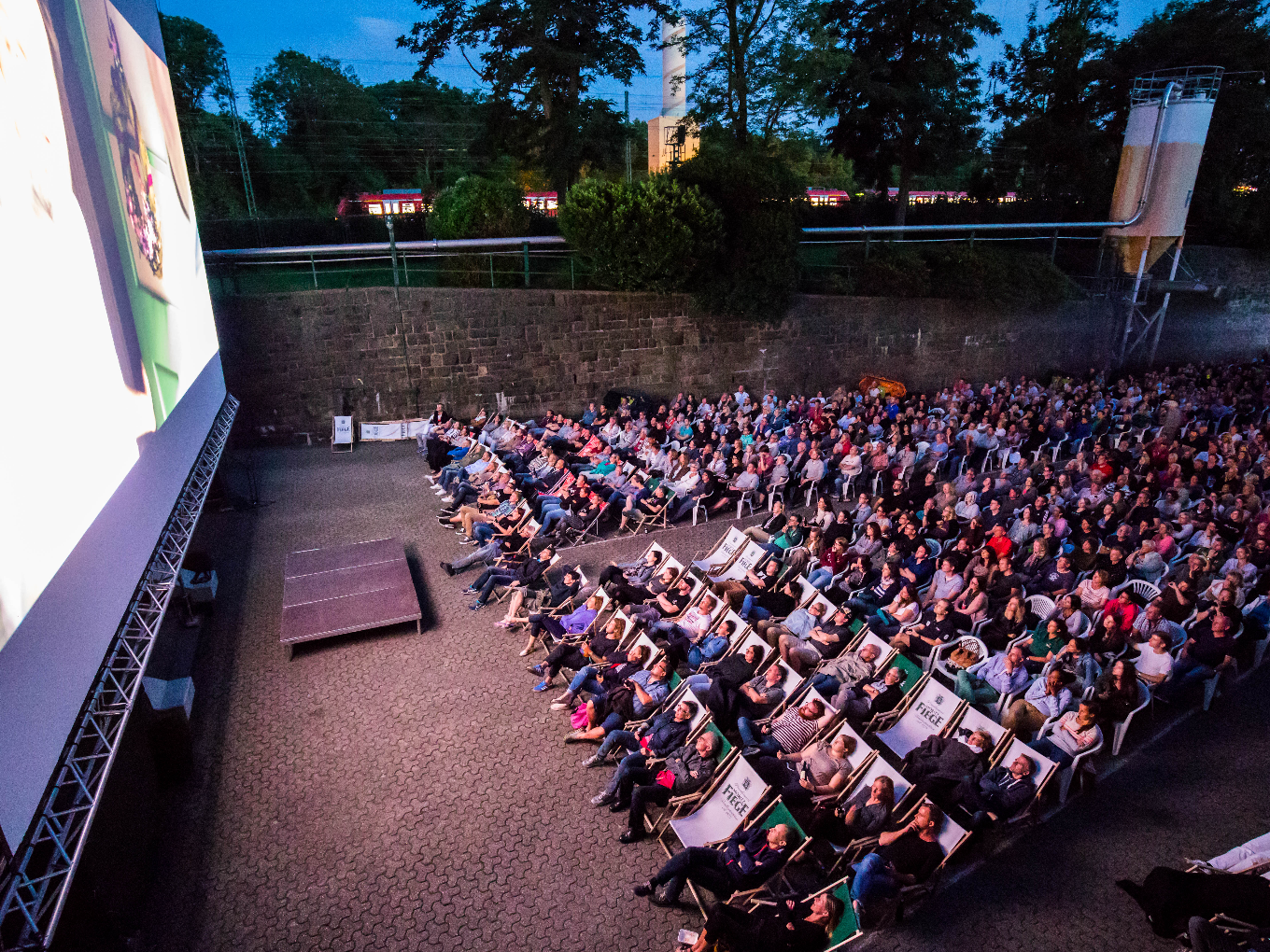 Großes Open-Air-Kino bei Abenddämmerung, viele Zuschauer auf Liegestühlen und Stühlen vor einer beleuchteten Leinwand.