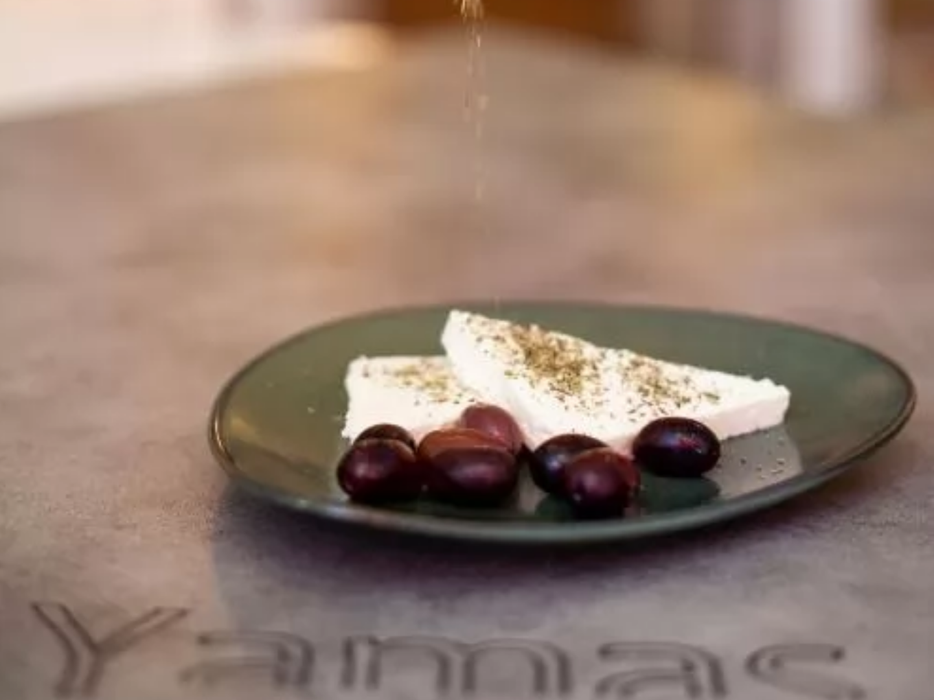 Zwei Stücke Feta-Käse mit Kräutern und mehrere schwarze Oliven auf einem grünen Teller, im Vordergrund der Schriftzug „Yamas“ auf dem Tisch.