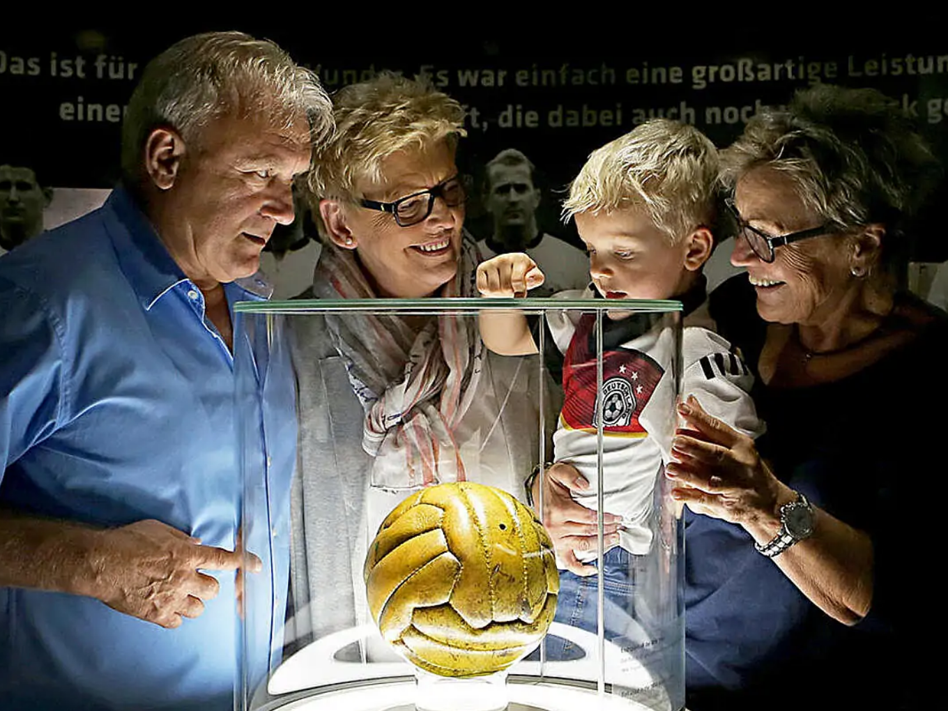 Vier Menschen, darunter ein Kind, betrachten einen goldenen Fußball in einer Glasvitrine.