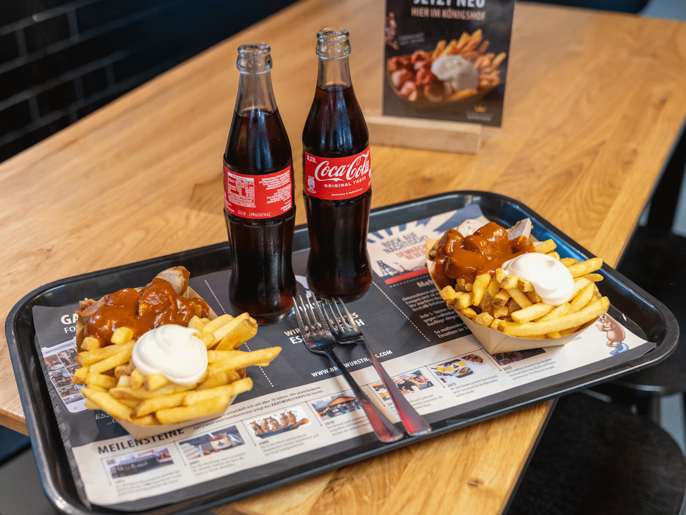 Zwei Portionen Currywurst mit Pommes und Sauce, zwei Flaschen Coca-Cola auf Tablett mit Speisekarte