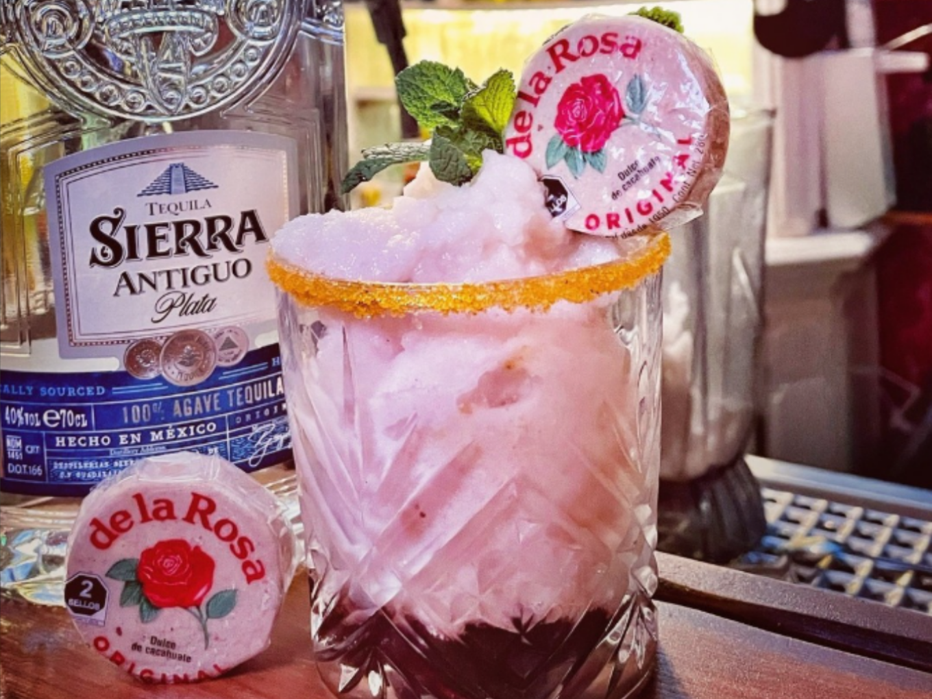 Rosa Cocktail mit Eis, Minze und einer „de la Rosa“-Süßigkeit, daneben eine Tequila-Flasche.