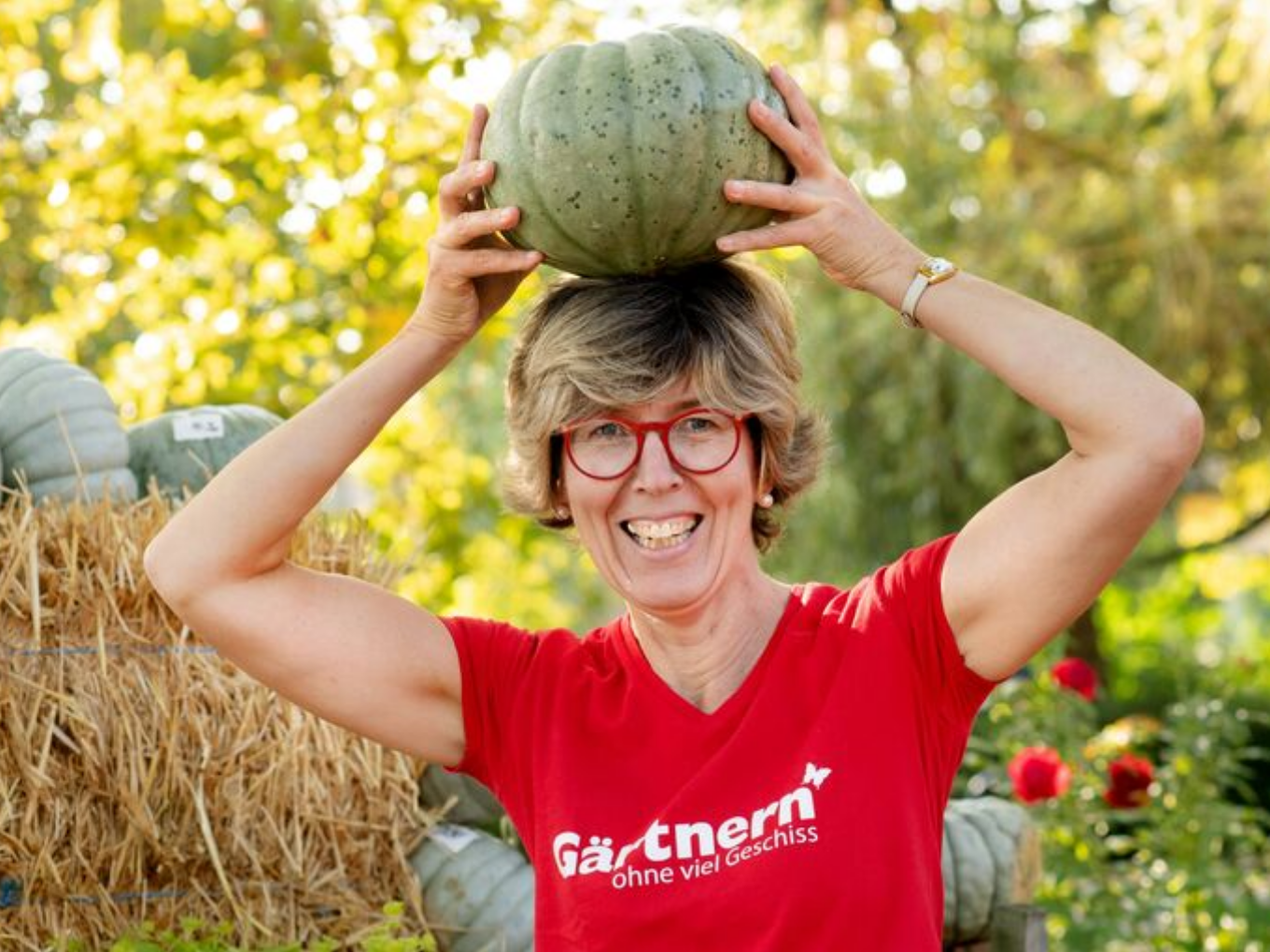 Frau mit Brille und rotem T-Shirt hält einen Kürbis lachend auf dem Kopf, im Hintergrund Strohballen und Garten.