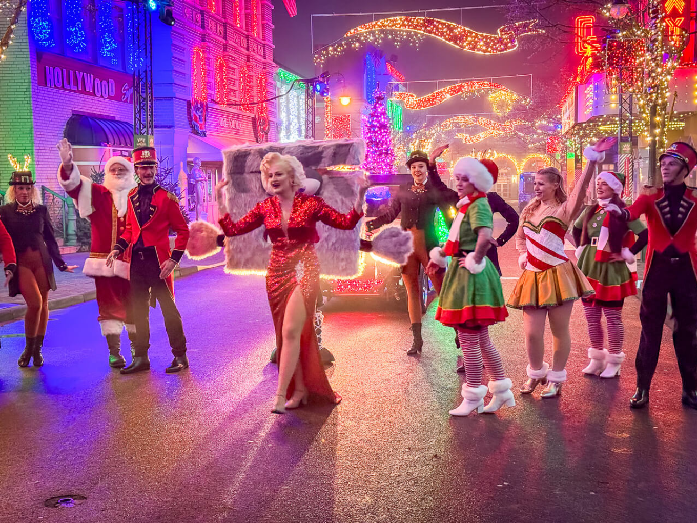 Weihnachtsparade mit Menschen in festlichen Kostümen und Lichterketten, eine Frau im roten Kleid steht im Mittelpunkt.