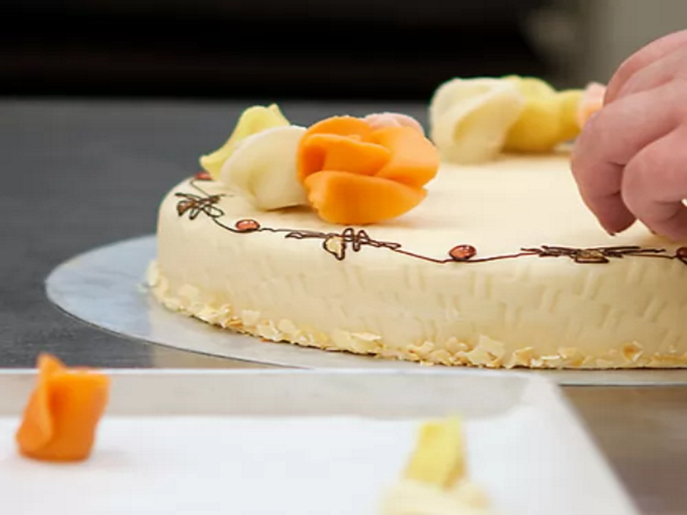 Eine Hand dekoriert eine Torte mit Marzipanblumen, die Torte ist mit heller Creme überzogen und auf einem silbernen Tablett platziert.