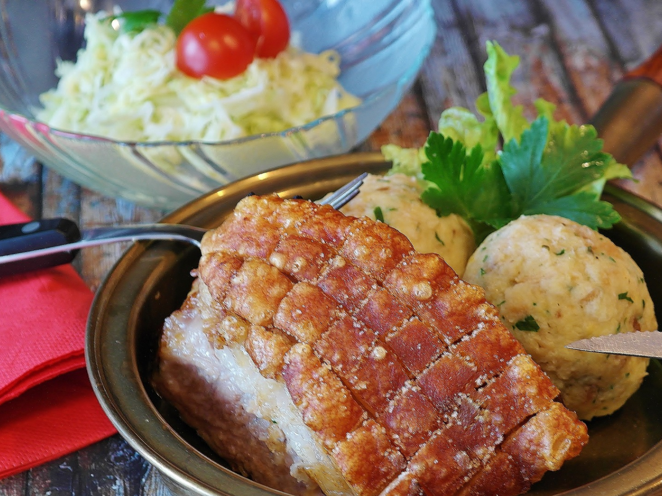 Teller mit knusprigem Schweinebraten, zwei Semmelknödeln und Salat im Hintergrund.
