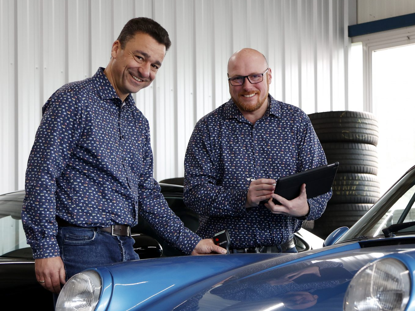 Zwei Männer in blauen Hemden stehen an einem blauen Auto und lächeln in die Kamera.