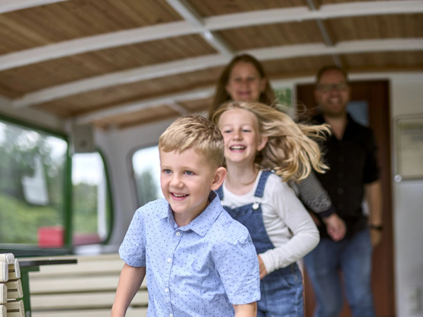 Zwei Kinder laufen lachend durch das Innere eines Ausflugsschiffs, im Hintergrund zwei Erwachsene.