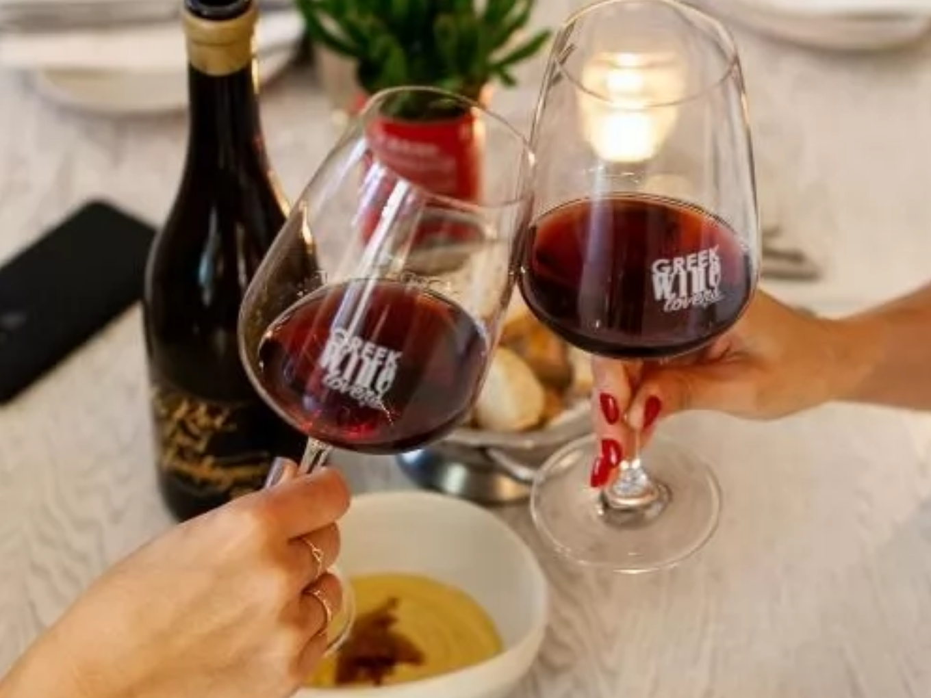 Zwei Personen stoßen mit Rotweingläsern an, auf den Gläsern steht „Greek Wild Lovers“, im Hintergrund eine Flasche Wein und ein gedeckter Tisch.