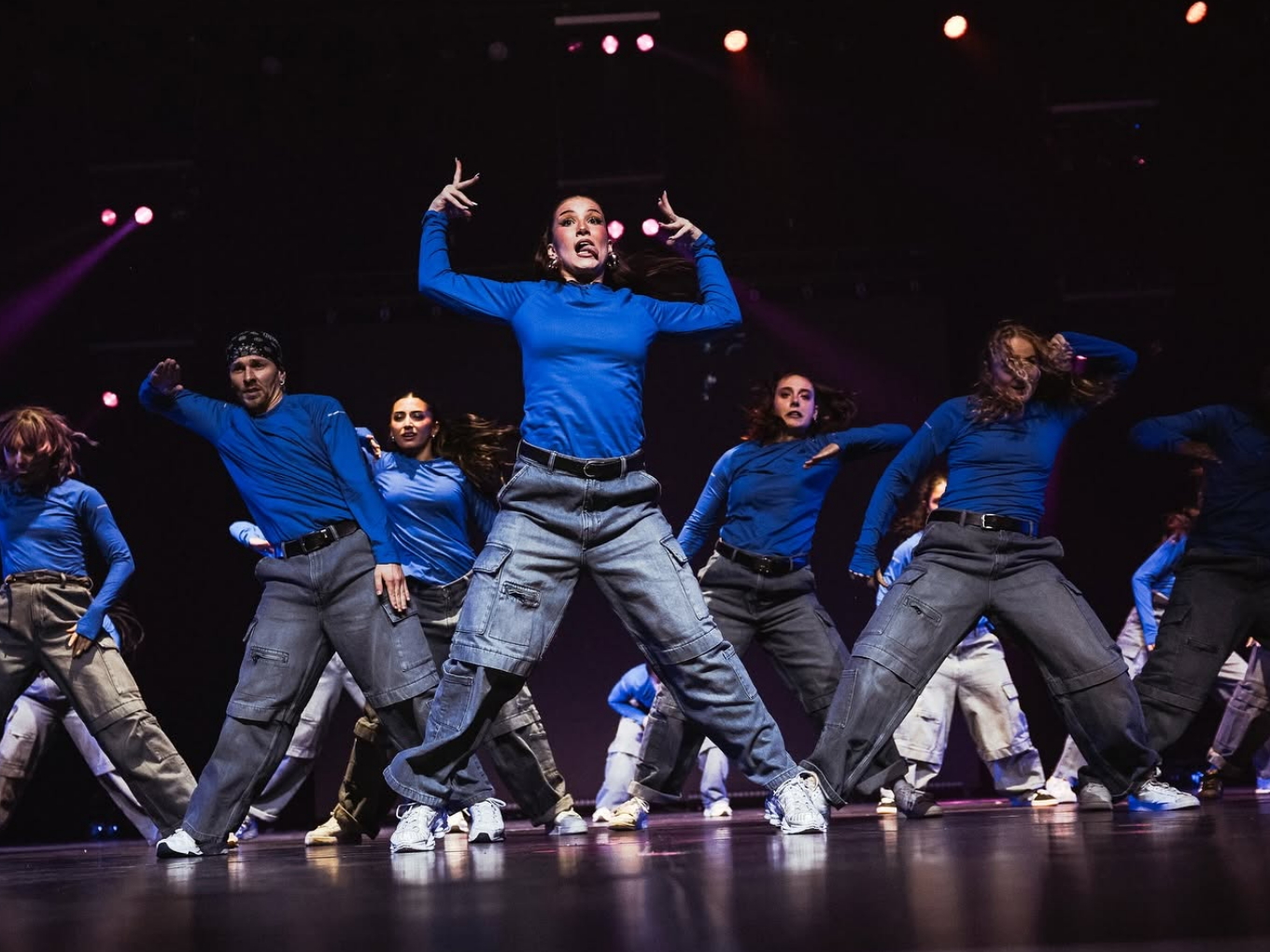 Tanzgruppe in blauen Oberteilen und grauen Hosen tanzt synchron auf der Bühne.