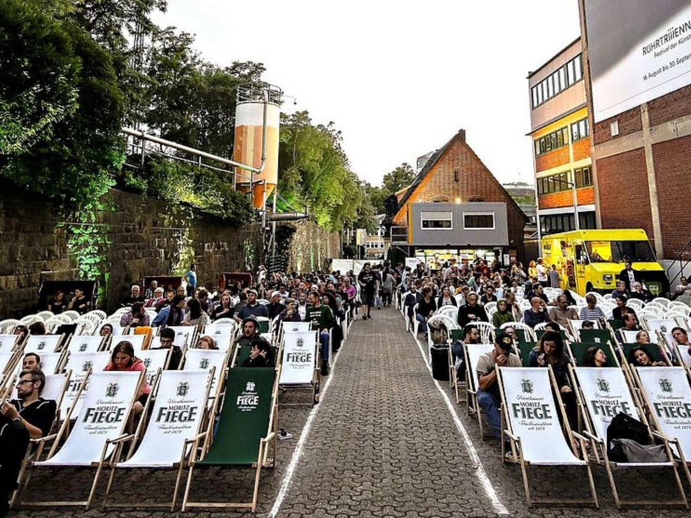 Open-Air-Kino mit zahlreichen weißen und grünen Fiege-Liegestühlen, viele Besucher und ein gelber Foodtruck im Hintergrund.