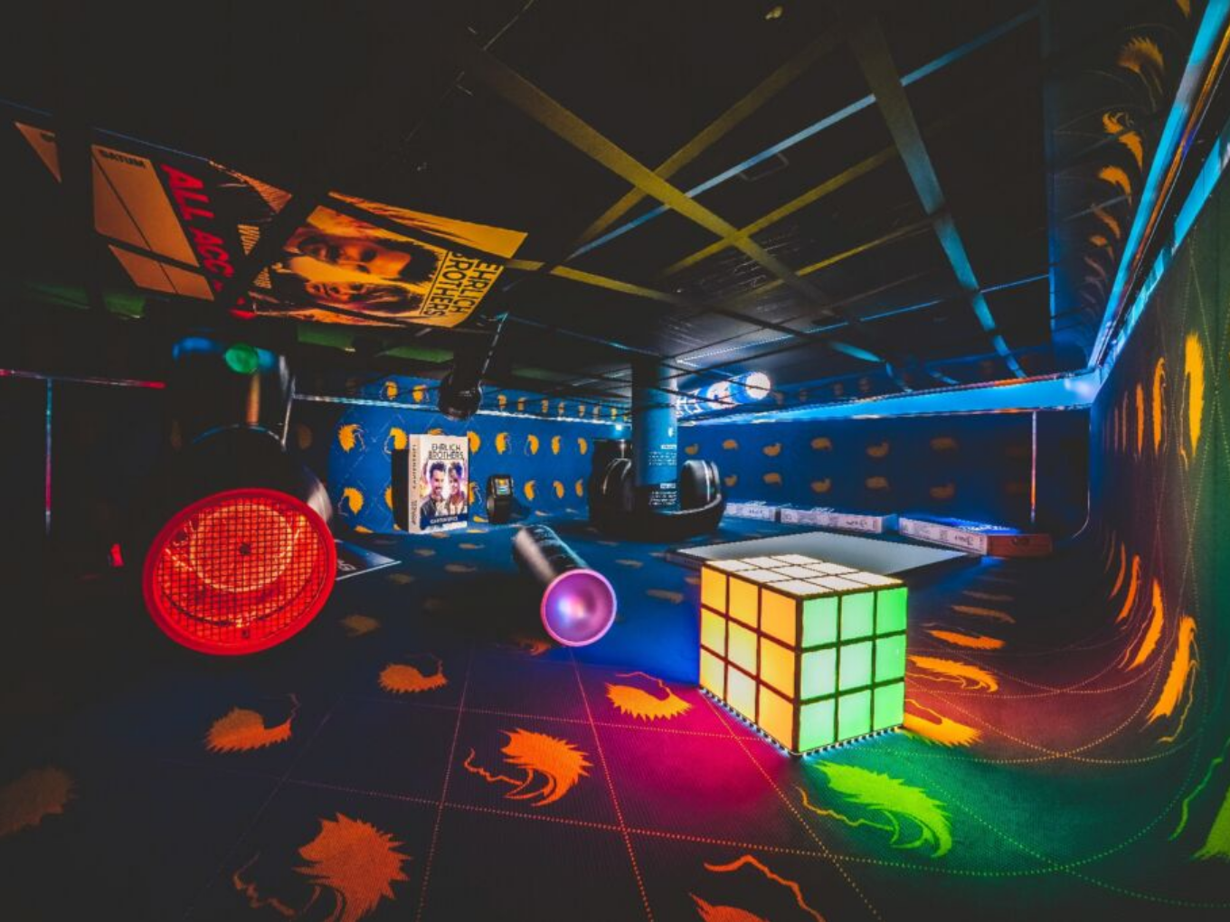 Ein Raum mit übergroßen Alltagsgegenständen wie einem Rubik’s Cube und einer Taschenlampe, alles bunt beleuchtet.