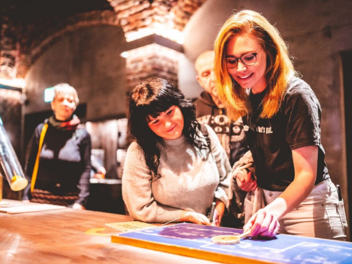 Zwei junge Frauen experimentieren an einem Tisch, andere Menschen beobachten sie im Hintergrund.