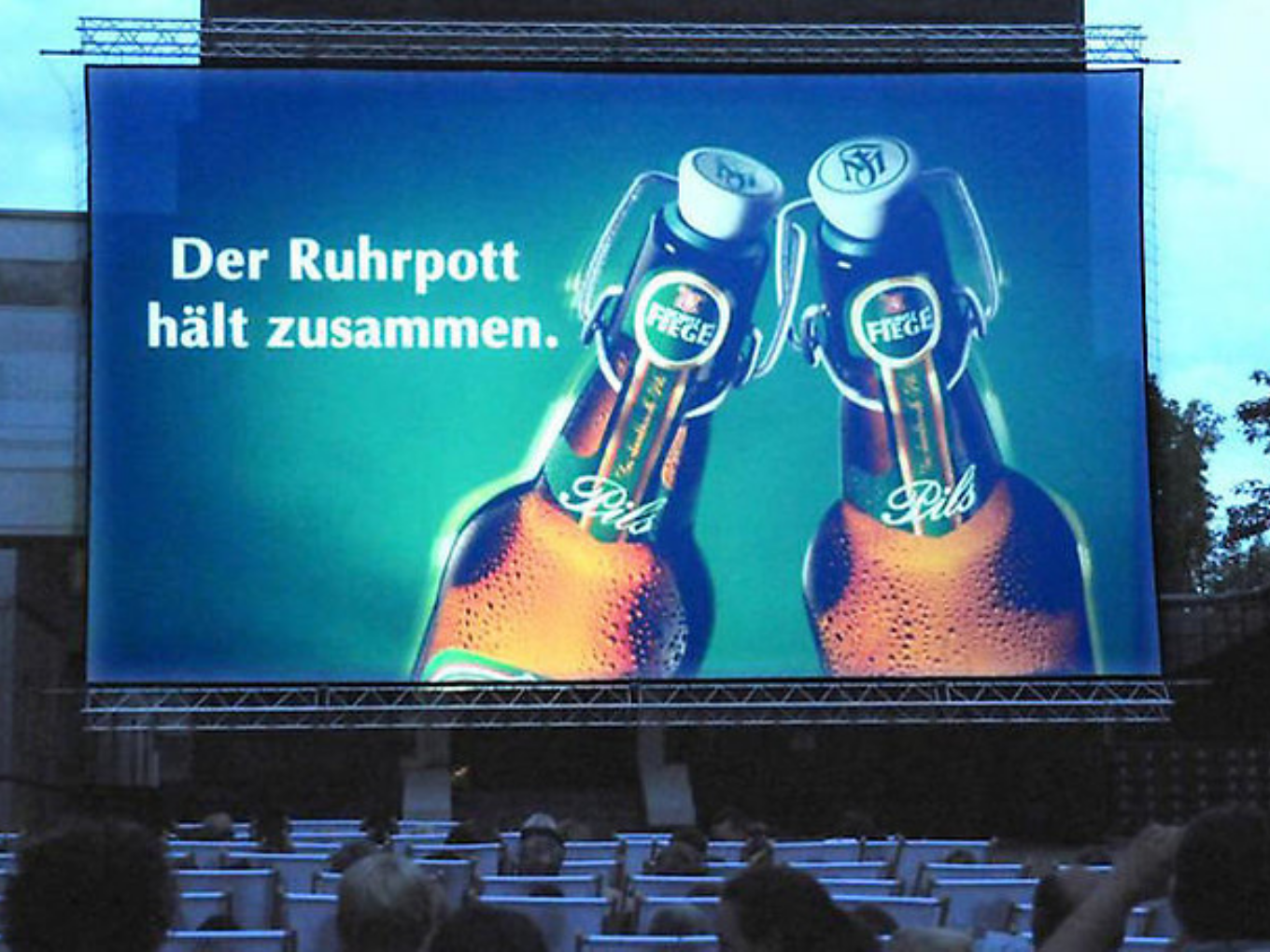 Open-Air-Kino mit großem Bildschirm, auf dem zwei Fiege-Bierflaschen und der Slogan „Der Ruhrpott hält zusammen“ zu sehen sind.