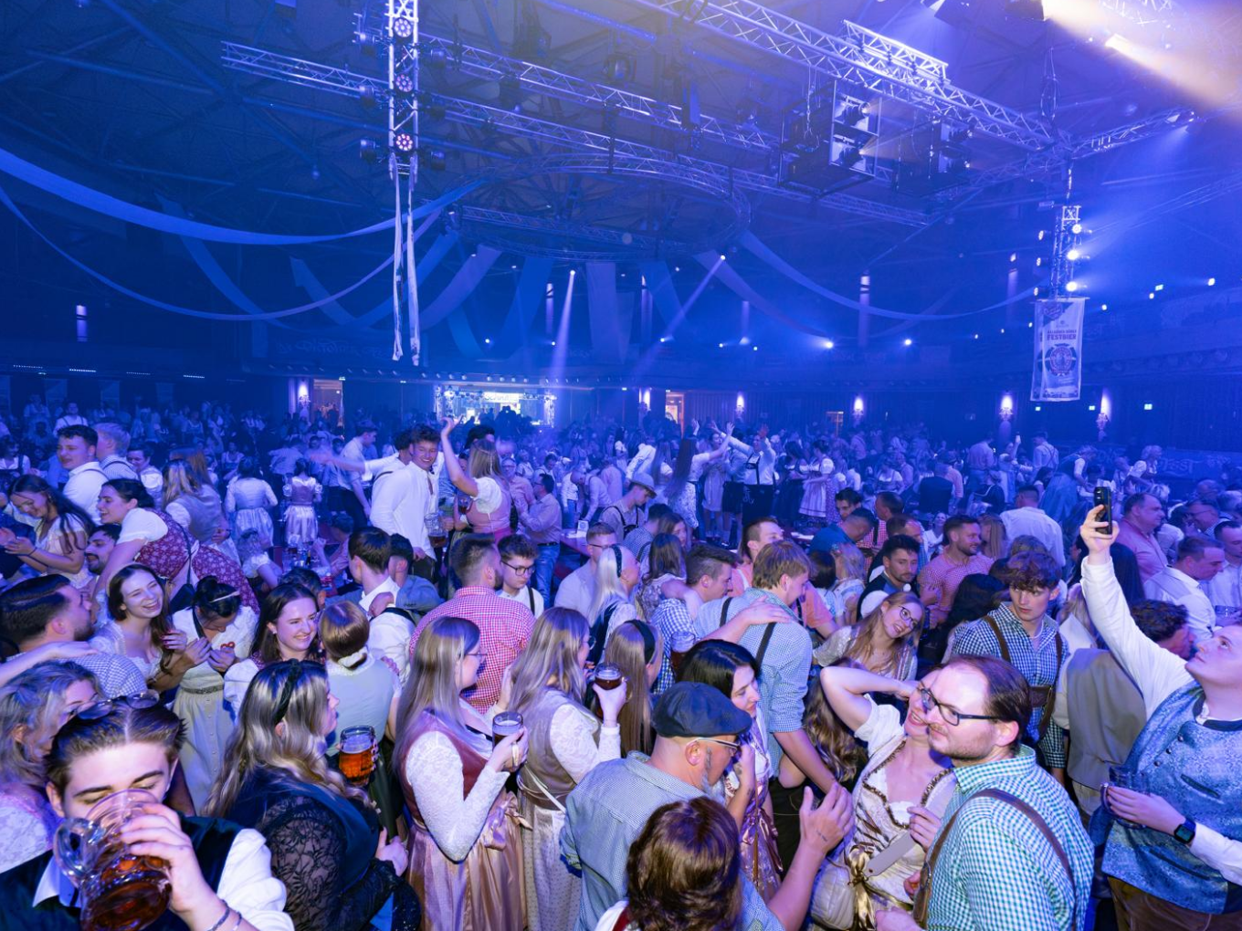 Menschen in Tracht feiern beim Oktoberfest Hagen in der blau beleuchteten Stadthalle.