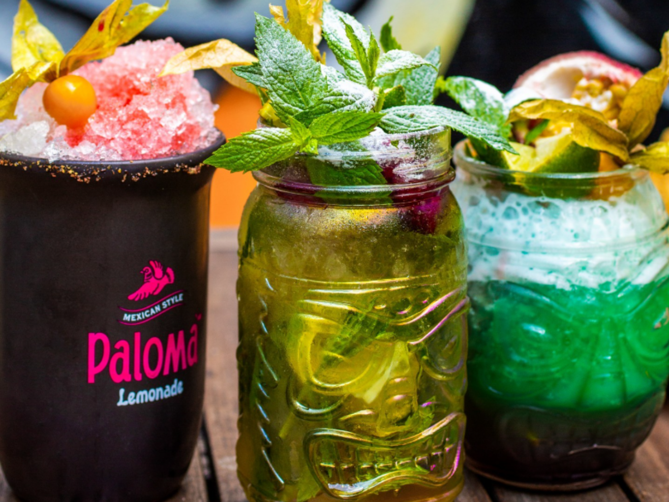 Drei bunte Cocktails in Gläsern mit Minze und Früchten, einer davon mit „Paloma Lemonade“-Aufdruck.