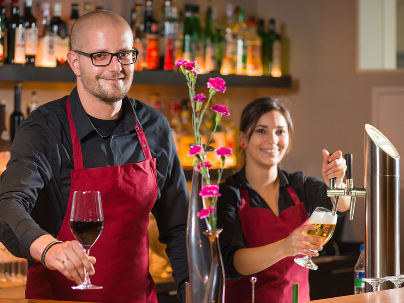 Zwei Servicekräfte hinter einer Bar, eine Person schenkt Bier ein, die andere hält ein Glas Rotwein.