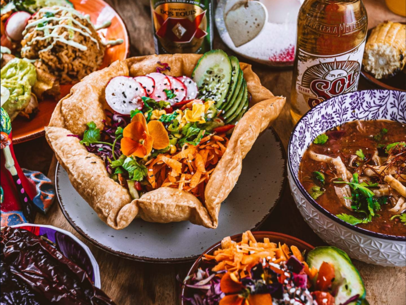 Bunte mexikanische Gerichte, darunter ein Taco-Salat in einer essbaren Schale, Suppe und Bierflaschen.