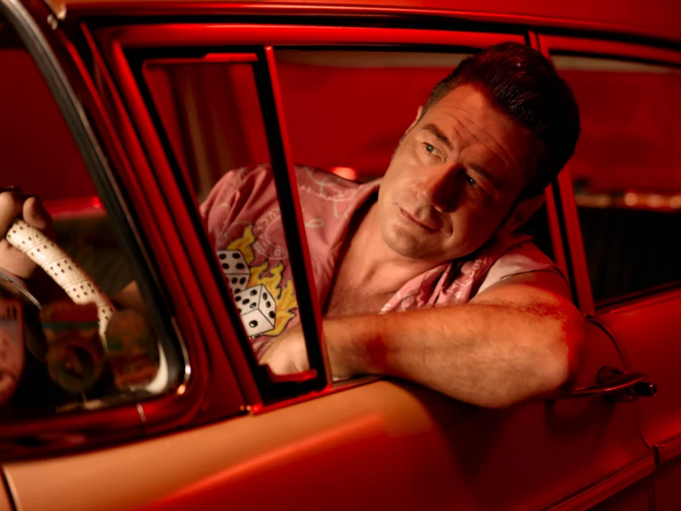 Mann mit Elvis-Frisur sitzt in einem roten Oldtimer und schaut aus dem Fenster.