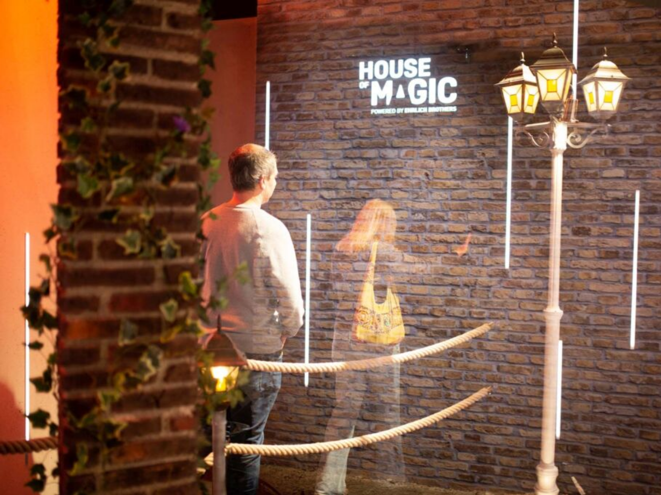 Ein Mann steht vor einer Wand mit dem Schriftzug „House of Magic“, daneben eine Laterne, eine Frau erscheint als durchsichtige Gestalt.