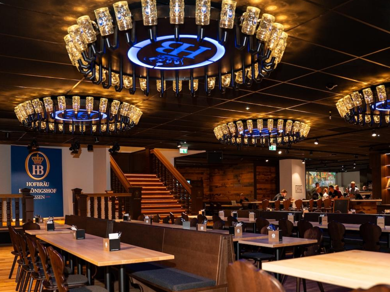 Großer Gastraum mit langen Holztischen und Kronleuchtern im Hofbräuhaus-Stil.