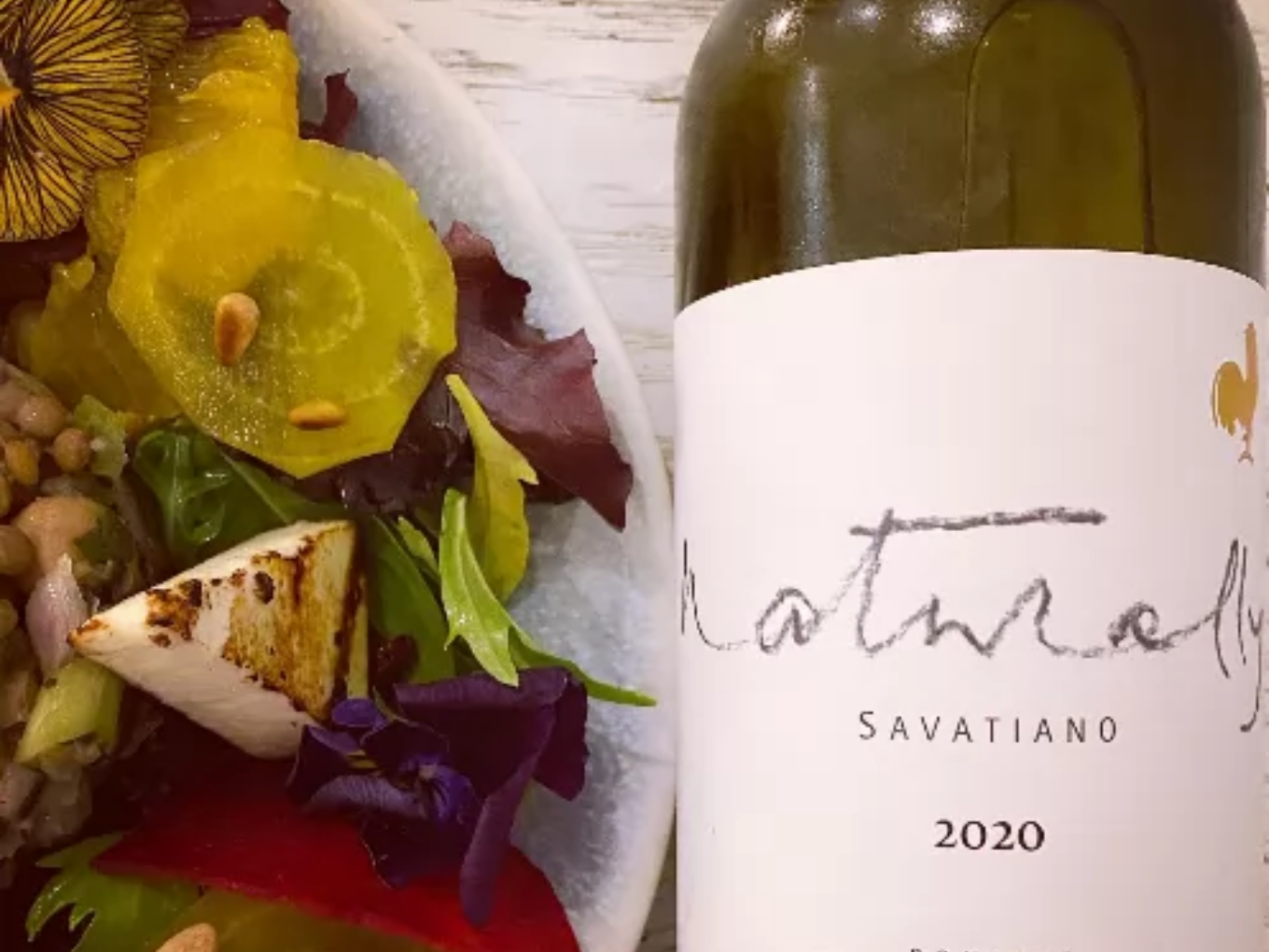 Ein Teller mit buntem Salat, gelben Rote-Bete-Scheiben, gegrilltem Käse und essbaren Blüten, daneben eine Flasche Weißwein mit dem Etikett „Savatiano 2020“.