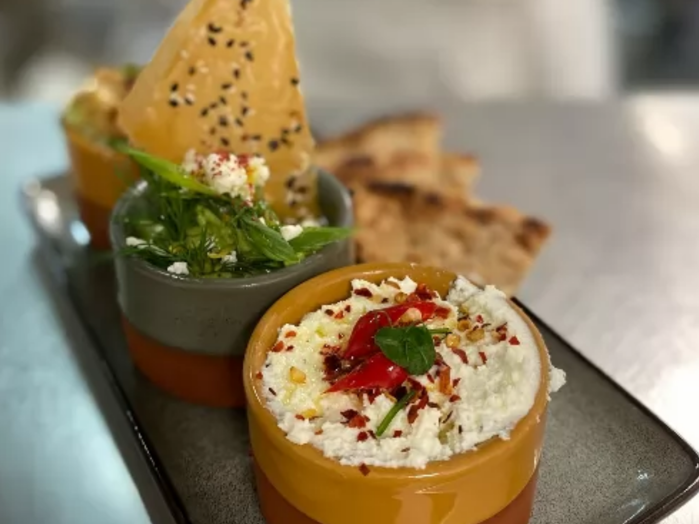 Drei kleine Schälchen mit griechischen Vorspeisen, darunter ein Dip mit Chili und Kräutern, ein grüner Salat und ein knuspriges Gebäckstück, im Hintergrund Fladenbrot.