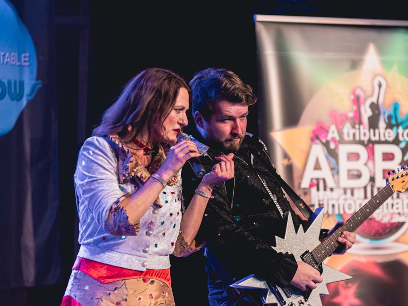 Nahaufnahme einer Sängerin und eines Gitarristen auf der Bühne während einer ABBA-Tribute-Show. Die Sängerin hält ein Mikrofon und singt, während der Gitarrist mit einer auffälligen, sternförmigen Gitarre spielt.