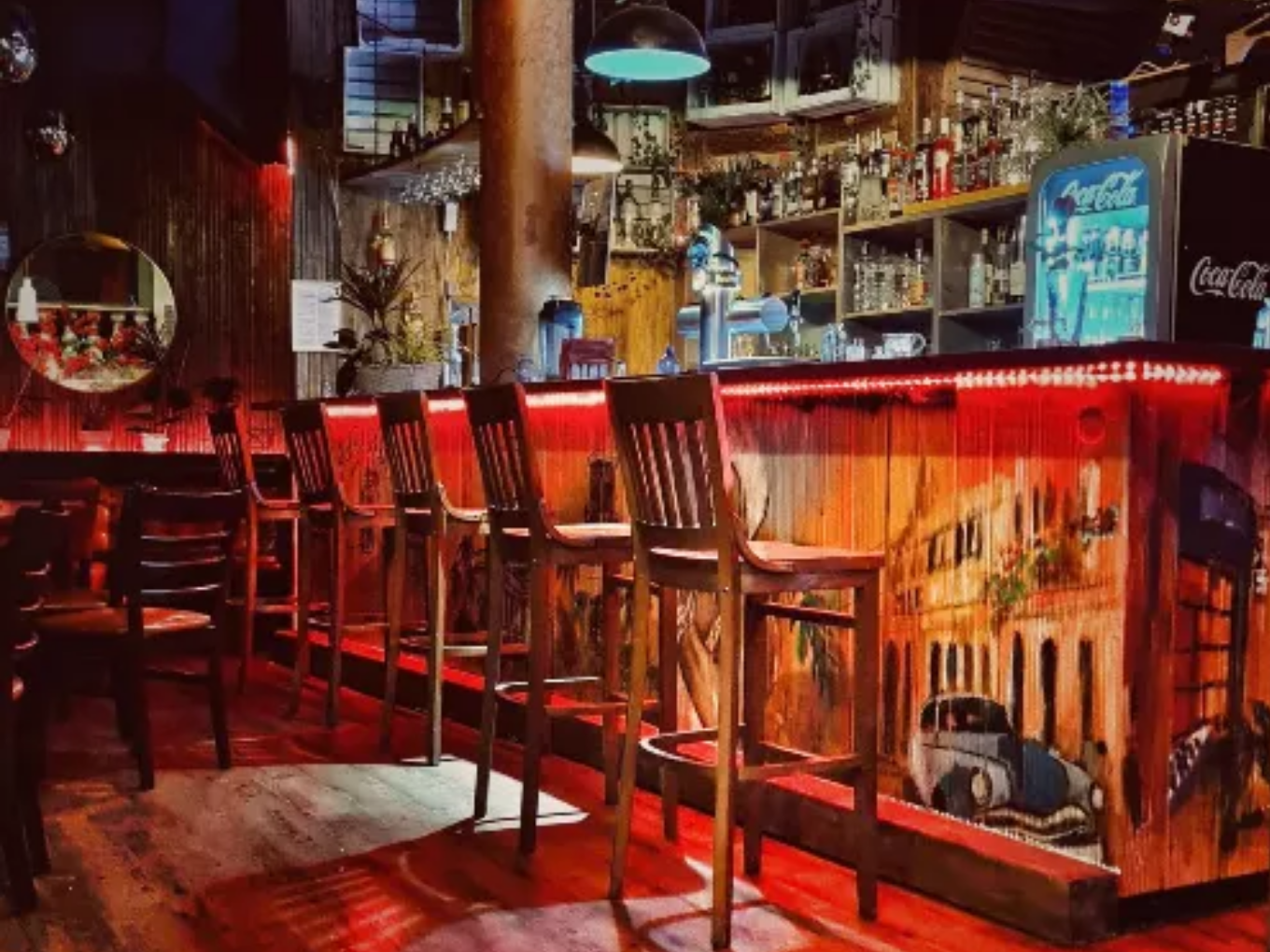 Eine stilvoll beleuchtete Bar mit hohen Holzstühlen, vielen Flaschen und Gläsern im Regal, sowie einer Theke mit einem Wandgemälde, das eine Straßenszene zeigt.