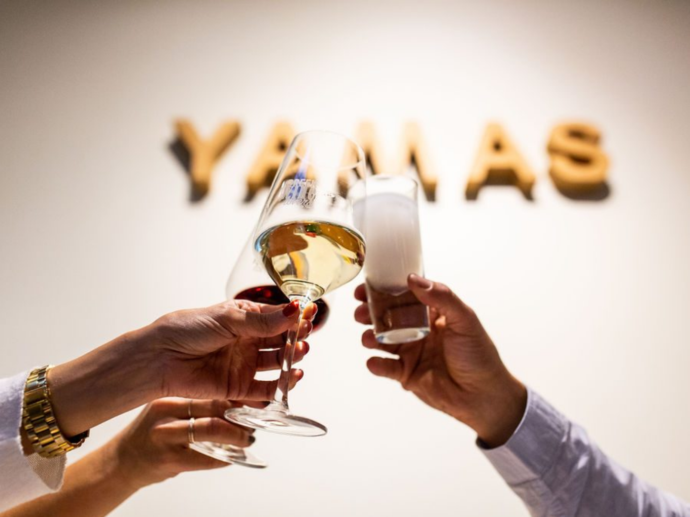 Drei Hände stoßen mit verschiedenen Gläsern an, im Hintergrund das Wort „YAMAS“ an der Wand.