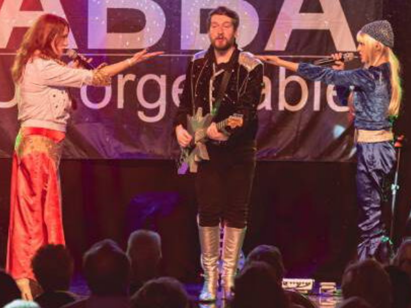 Drei Musiker auf einer Bühne bei einer ABBA-Tribute-Show. Zwei Frauen stehen links und rechts mit Mikrofonen und singen, während ein Mann in der Mitte eine E-Gitarre spielt.