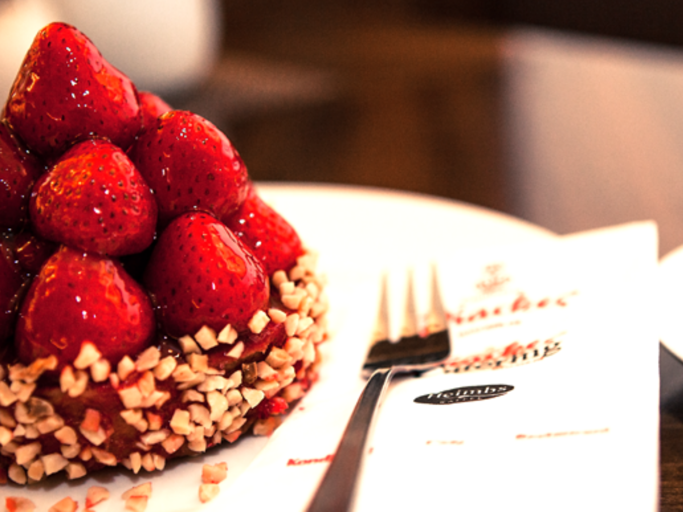 Erdbeerkuchen mit frischen, glänzenden Erdbeeren und gehackten Nüssen am Rand, auf einem Teller mit Gabel und Serviette.