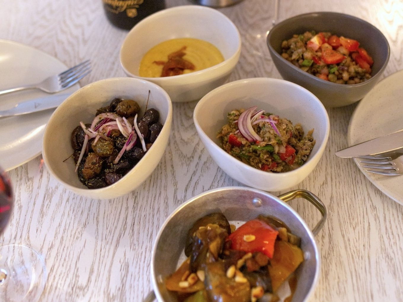 Verschiedene griechische Vorspeisen in kleinen Schalen, darunter Oliven mit Zwiebeln, Hummus, Linsensalat und geschmorte Auberginen, auf einem weiß gedeckten Tisch.
